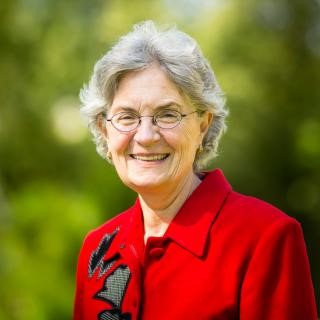Sherrie Brown, a woman with gray hair smiling with glasses and wearing a red jacket with black standing outside against a green background.  